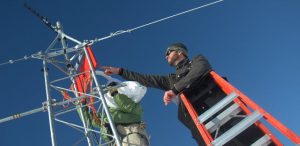 Two people work on a metal weather station tower in Antarctica; one stands on an orange ladder adjusting equipment, both wearing winter jackets and hats under a clear blue sky during an Antarctic Logistics and Expeditions mission.