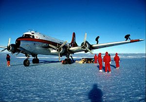 A vintage propeller plane is parked on snowy ground under a clear blue sky in Antarctica. Several people in red cold-weather gear, part of Antarctic Logistics and Expeditions, stand and work near the aircraft with supplies and equipment.