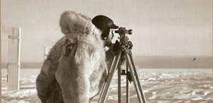 A person wearing a heavy fur coat and hood looks through a surveying instrument on a tripod, standing on Antarctica's snow-covered landscape under a cloudy sky—part of Antarctic Logistics and Expeditions' challenging fieldwork.