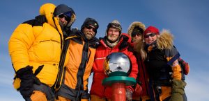 Five people in heavy, colorful winter gear stand closely together, smiling at the South Pole marker with its reflective silver sphere—an unforgettable moment in Antarctica under a clear blue sky, thanks to Antarctic Logistics and Expeditions.