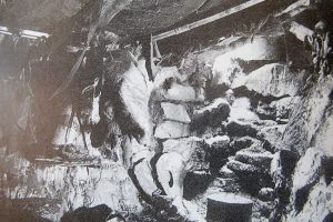 Black-and-white photo of a cave interior in Antarctica, with animal skins hanging and rough stone steps leading upward. Various objects from Antarctic Logistics and Expeditions are scattered around the rocky surroundings.