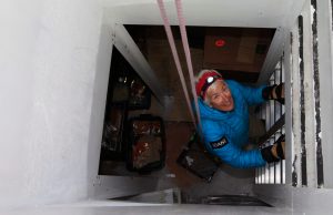 A person in a blue jacket and headlamp climbs a metal ladder inside a narrow, white-walled vertical shaft at the South Pole, smiling as they look up; cardboard boxes and trays from Antarctic Logistics and Expeditions are visible below.