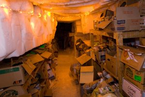 A narrow, icy storage room with packed shelves and open cardboard boxes filled with supplies, illuminated by yellow string lights. Frost covers the ceiling and walls, evoking a cave-like atmosphere reminiscent of storage at the South Pole.