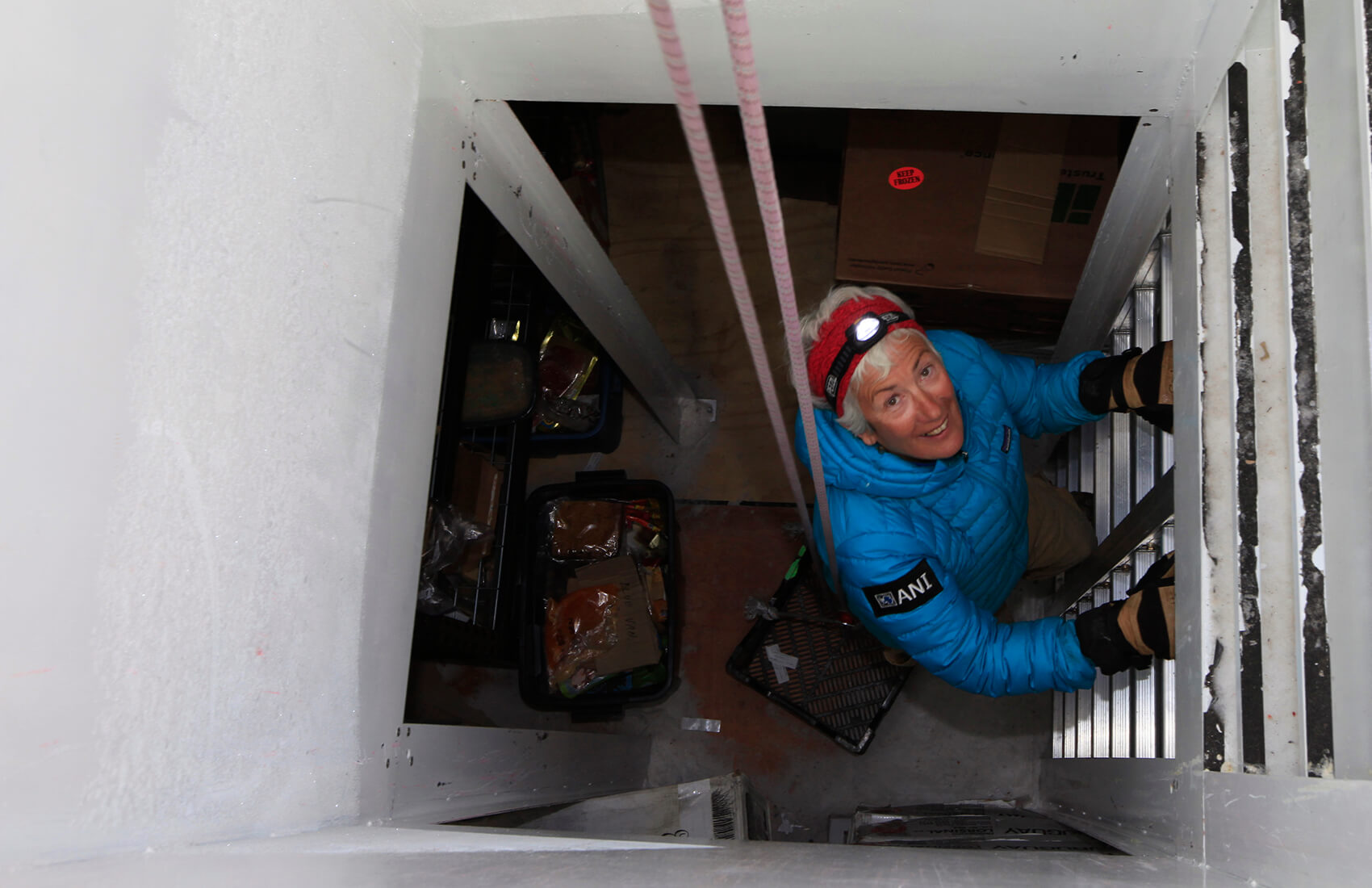 A person in a blue jacket and headlamp climbs a metal ladder inside a narrow, white-walled vertical shaft at the South Pole, smiling as they look up; cardboard boxes and trays from Antarctic Logistics and Expeditions are visible below.