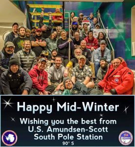 A group of people pose together on indoor stairs in Antarctica, smiling in their winter clothing. Below them, a sign reads: Happy Mid-Winter. Wishing you the best from U.S. Amundsen-Scott South Pole Station 90°S.