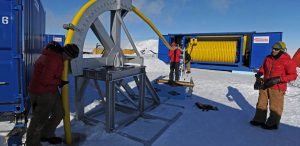 Three people in red jackets work with large yellow hoses and metal equipment on the snowy landscape of Antarctica, with blue storage containers and machinery from Antarctic Logistics and Expeditions in the background under a clear sky.