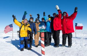 A group of people in heavy winter clothing stand on snowy ground at the South Pole, smiling and waving beside a striped pole topped with a sphere. American and Chilean flags are visible in the background under Antarctica's clear blue sky.