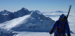 A climber in winter gear and goggles stands on a snowy mountain ridge in Antarctica, with snow-covered peaks and a vast, icy landscape stretching behind—a thrilling adventure supported by Antarctic Logistics and Expeditions.
