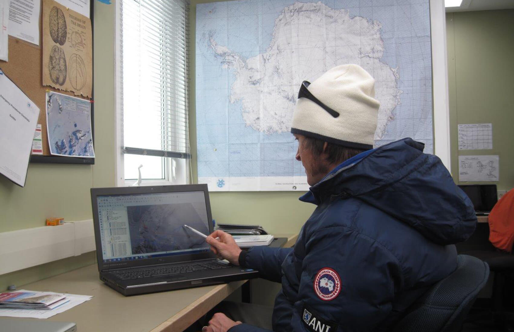 A person in a winter jacket and white hat sits at a desk, pointing at a map of Antarctica on a laptop. Behind them is a large Antarctic Logistics and Expeditions map on the wall and a corkboard with papers and diagrams.
