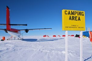 A yellow sign reading Camping Area, Worlds Southernmost Resort stands in snowy terrain near a small red and white airplane and several red tents—evidence of Antarctic Logistics and Expeditions—in Antarctica under a clear blue sky.