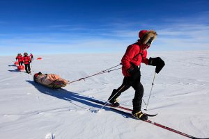 A group of people in red jackets and cold-weather gear pull sleds across a snowy, flat landscape under a clear blue sky, evoking an Antarctic Logistics and Expeditions journey across Antarctica.