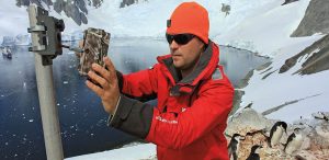 A person in a red jacket and orange hat sets up a camera on a pole in snowy Antarctica near water, with penguins standing nearby—capturing the essence of an Antarctic Logistics and Expeditions adventure.