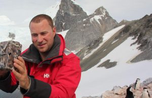A man in a red jacket sets up a camouflaged camera on a rocky, snowy mountain in Antarctica, with two penguins standing nearby. Antarctic Logistics and Expeditions support is evident amid the snow-covered peaks in the background.