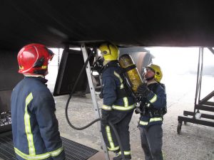 Three firefighters in protective gear and helmets work together in Antarctica; one climbs a ladder with a hose under a large structure while the others assist and observe, showcasing teamwork in challenging South Pole conditions.