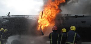 Firefighters in helmets and protective gear spray water on a large fire engulfing the side of an aircraft during a daytime Antarctic Logistics and Expeditions emergency response or training exercise. Thick flames and smoke are visible.