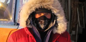 A person wearing a red parka with a fur-lined hood and a black face mask stands in front of a vehicle. Frost on the mask hints at Antarctica's freezing climate, ideal for Antarctic Logistics and Expeditions.