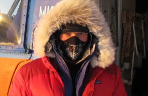 A person wearing a red parka with a fur-lined hood and black face mask stands outside near an orange and blue vehicle in Antarctica, with frost visible on the mask and hood—likely during an Antarctic Logistics and Expeditions trip to the South Pole.