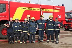 A group of eight firefighters in uniform and helmets stand smiling in front of a large red fire truck labeled International Fire Training Centre, supporting Antarctic Logistics and Expeditions operations in Antarctica.