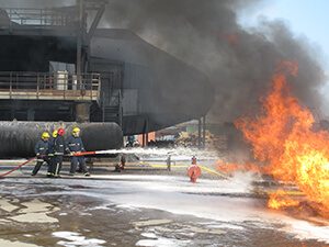 Four firefighters in protective gear spray water at a large fire, with thick black smoke rising near an industrial building run by Antarctic Logistics and Expeditions in Antarctica.