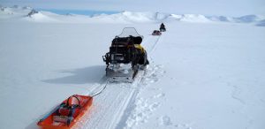 Two snowmobiles towing sleds travel across a vast, snowy landscape in Antarctica, with mountains in the distance under a cloudy sky. The snowmobiles, part of Antarctic Logistics and Expeditions, leave clear tracks in the deep snow.