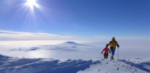 Two people in winter gear hike on a snowy mountain under a bright sun, exploring an Antarctic landscape with vast, snow-covered terrain stretching beneath a clear blue sky—an adventure made possible by Antarctic Logistics and Expeditions.