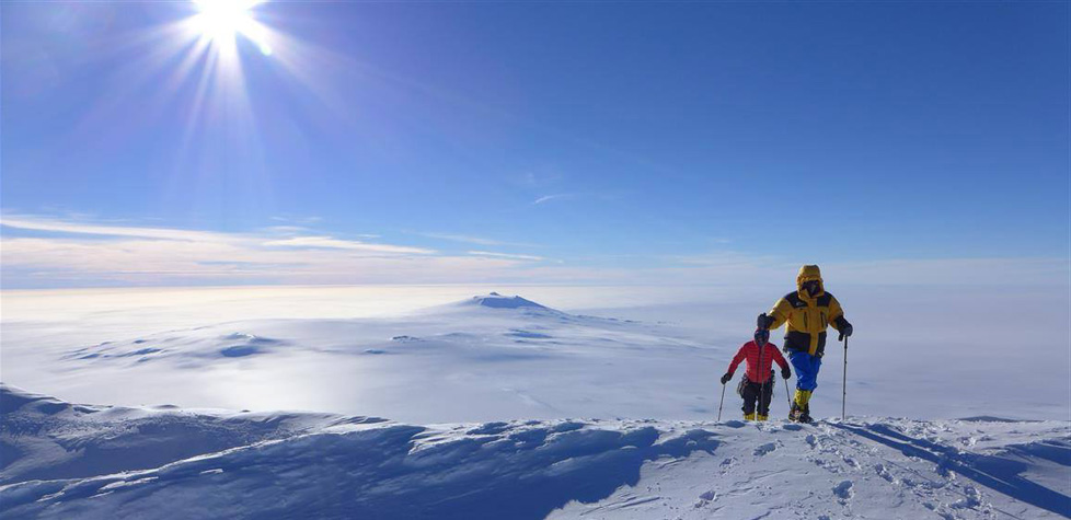 Two people in winter gear hike on a snowy mountain under a bright sun, exploring an Antarctic landscape with vast, snow-covered terrain stretching beneath a clear blue sky—an adventure made possible by Antarctic Logistics and Expeditions.