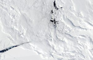 A satellite view of Antarctica's vast icy landscape reveals cracks, textured snow, and dark patches of exposed water or ice near the center, capturing the rugged, frozen environment of the South Pole.