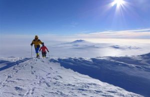 Two people in winter gear hike on a snowy mountain ridge under a bright sun, with vast snow-covered landscape and distant peaks of Antarctica visible—an adventure reminiscent of Antarctic Logistics and Expeditions en route to the South Pole.
