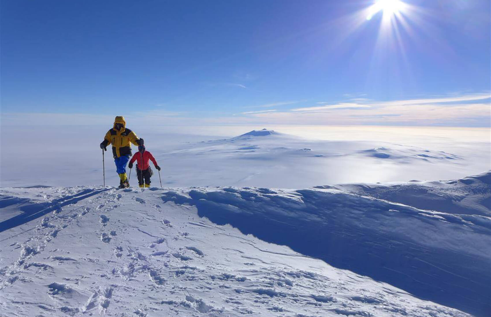Two people in winter gear hike on a snowy mountain ridge under a bright sun, with vast snow-covered landscape and distant peaks of Antarctica visible—an adventure reminiscent of Antarctic Logistics and Expeditions en route to the South Pole.
