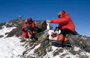 Two people in heavy red parkas and snow boots sit on rocky, snow-covered terrain in Antarctica with laptops and scientific equipment, working outdoors under a clear blue sky as part of an Antarctic Logistics and Expeditions mission.