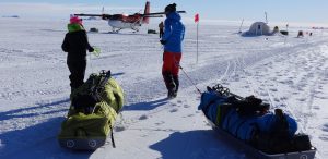 Two people in winter gear pull sleds loaded with gear across snowy Antarctic terrain toward a small airplane and camp setup in the distance, under a clear blue sky—showcasing the spirit of Antarctic Logistics and Expeditions.