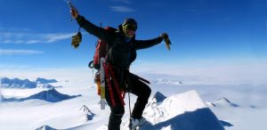 A climber wearing winter gear and sunglasses stands triumphantly on a snowy Antarctic peak with arms raised, surrounded by a panoramic view of Antarctica’s snow-covered summits under a clear blue sky—an epic moment with Antarctic Logistics and Expeditions.