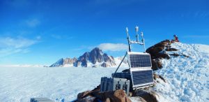 A weather station with solar panels sits on a snowy, rocky hilltop in Antarctica, with mountains and a vast ice field stretching into the distance under a clear blue sky.