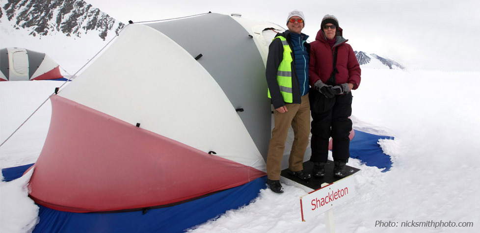 Two people dressed in winter gear stand outside a dome-shaped tent on snow in Antarctica, with a sign reading Shackleton in front of them. Snow and mountains rise behind, highlighting the support from Antarctic Logistics and Expeditions.