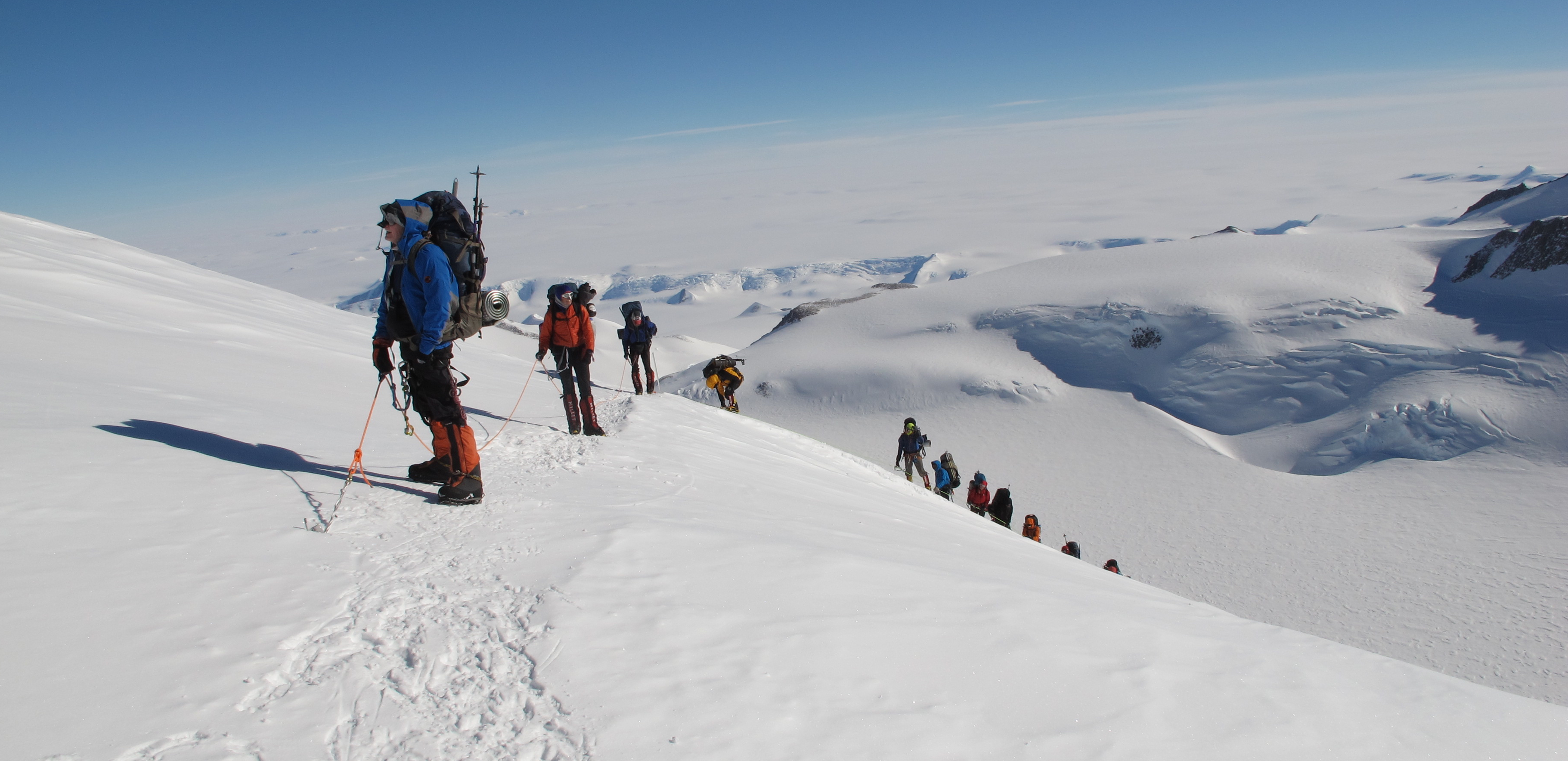 A group of climbers in winter gear hikes in a line along a snowy mountain ridge in Antarctica, surrounded by snow-covered peaks and a vast, white landscape under a clear blue sky—an adventure supported by Antarctic Logistics and Expeditions.