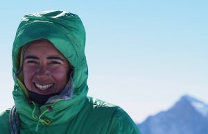 A person in a green hooded jacket smiles outdoors with an Antarctic mountain peak behind them, beneath a clear blue sky—a moment made possible by Antarctic Logistics and Expeditions.