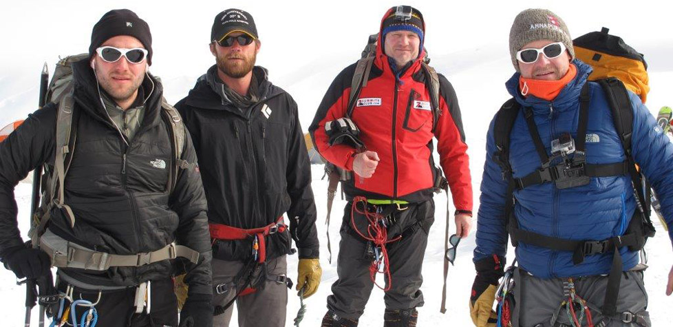 Four men wearing winter jackets, sunglasses, and climbing gear stand side by side on a snowy mountain in Antarctica, posing for a photo during their mountaineering expedition with Antarctic Logistics and Expeditions.