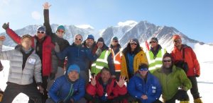A group of people in colorful winter gear pose, smiling and cheering, with snowy mountains and blue sky behind them. Some wear sunglasses and reflective vests, hinting at an Antarctic Logistics and Expeditions adventure in Antarctica.