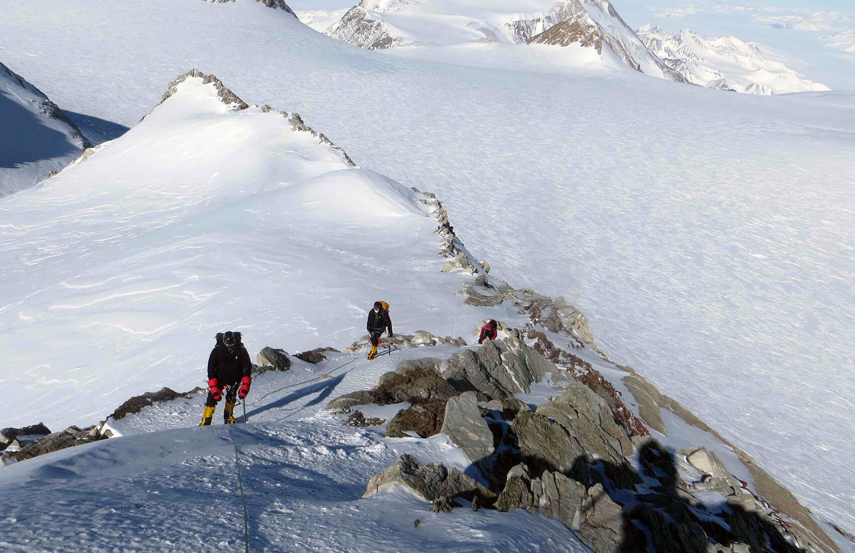 Three climbers in winter gear ascend a rocky, snow-covered mountain ridge in Antarctica, with a vast expanse of snow and distant peaks visible under a clear sky.