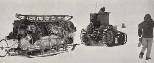 A tracked vehicle pulls a large sled loaded with supplies across a snowy landscape in Antarctica, while two people in heavy winter clothing walk nearby. The scene suggests Antarctic Logistics and Expeditions during a polar supply transport.