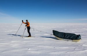 A person in an orange jacket skis across a vast snowy landscape in Antarctica, pulling a sled loaded with gear under the clear blue sky, reminiscent of South Pole expeditions supported by Antarctic Logistics and Expeditions.