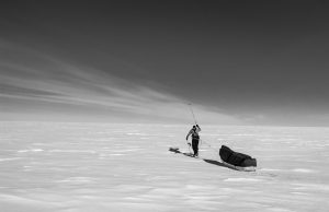 A person pulls a large sled across a vast, snowy landscape under a clear sky in Antarctica, casting a long shadow on the snow. The scene is in black and white.