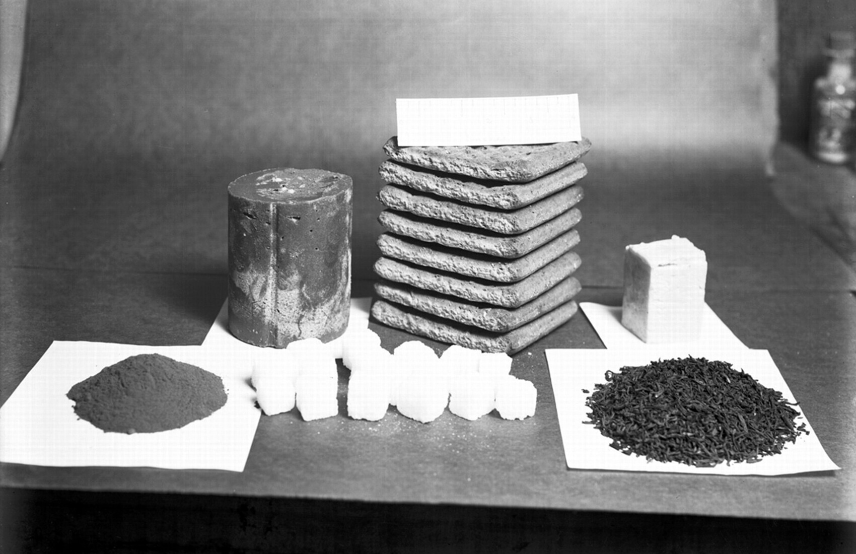 Black-and-white photo of various food rations on paper, including a stack of rectangular blocks, a round can, sugar cubes, and tea leaves—supplies reminiscent of what explorers once relied on during early South Pole expeditions in Antarctica.