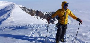 A person wearing a yellow jacket and snow gear stands on a snowy mountain ridge in Antarctica, holding two trekking poles, with snow-covered peaks and cliffs in the background under a clear sky—a true Antarctic Logistics and Expeditions moment.