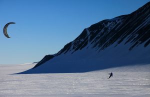 A person kite skiing on a snowy plain in Antarctica, beside a steep, shadowed mountain slope under a clear blue sky—a thrilling adventure made possible by Antarctic Logistics and Expeditions.
