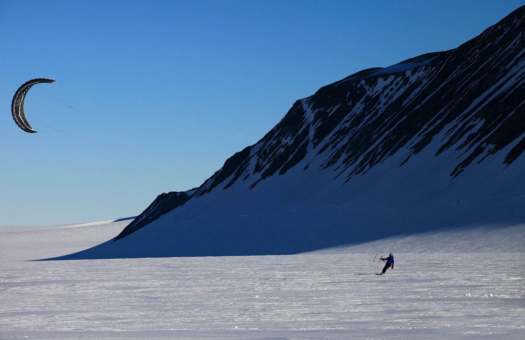 A person kite skiing on a snowy plain in Antarctica, beside a steep, shadowed mountain slope under a clear blue sky—a thrilling adventure made possible by Antarctic Logistics and Expeditions.