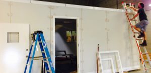 A person on a ladder works near the roof of a white, partially constructed room inside a larger building—typical of Antarctic Logistics and Expeditions sites. Another ladder and door frames lean against the wall by a doorway.