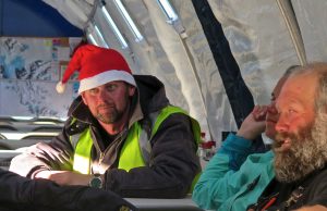 Two men sit inside a tent-like structure in Antarctica. One, wearing a Santa hat and high-visibility vest, looks toward the camera. The other, with a beard and teal jacket, smiles as maps from Antarctic Logistics and Expeditions hang behind them.