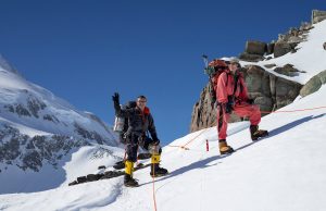Two climbers in winter gear stand on a snowy Antarctic slope, roped together, with one waving at the camera. Rocky cliffs and another snow-covered peak rise under a clear blue sky—an adventure with Antarctic Logistics and Expeditions.