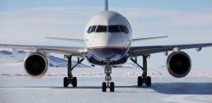 A large commercial airplane operated by Antarctic Logistics and Expeditions is parked on a snowy, icy runway, with snow-covered mountains and a cloudy Antarctic sky in the background. The photo is taken from directly in front of the aircraft.